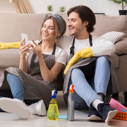 Couple sitting on floor in aprons and gloves, looking at a smartphone with cleaning supplies nearby.