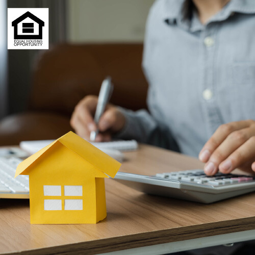 A small yellow paper house on a desk with a person using a calculator and pen in the background.
