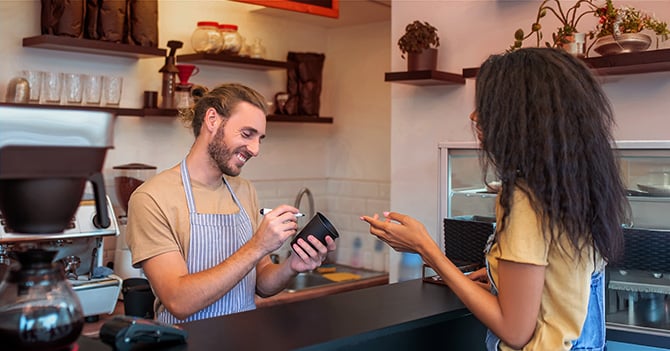 A barista writes down a coffee order on a cup while the customer orders.